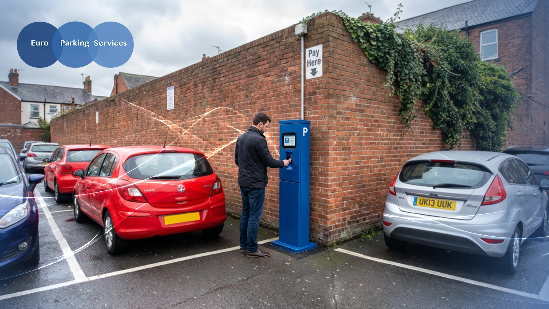 A man using a pay and display parking machine in a car park with parked cars