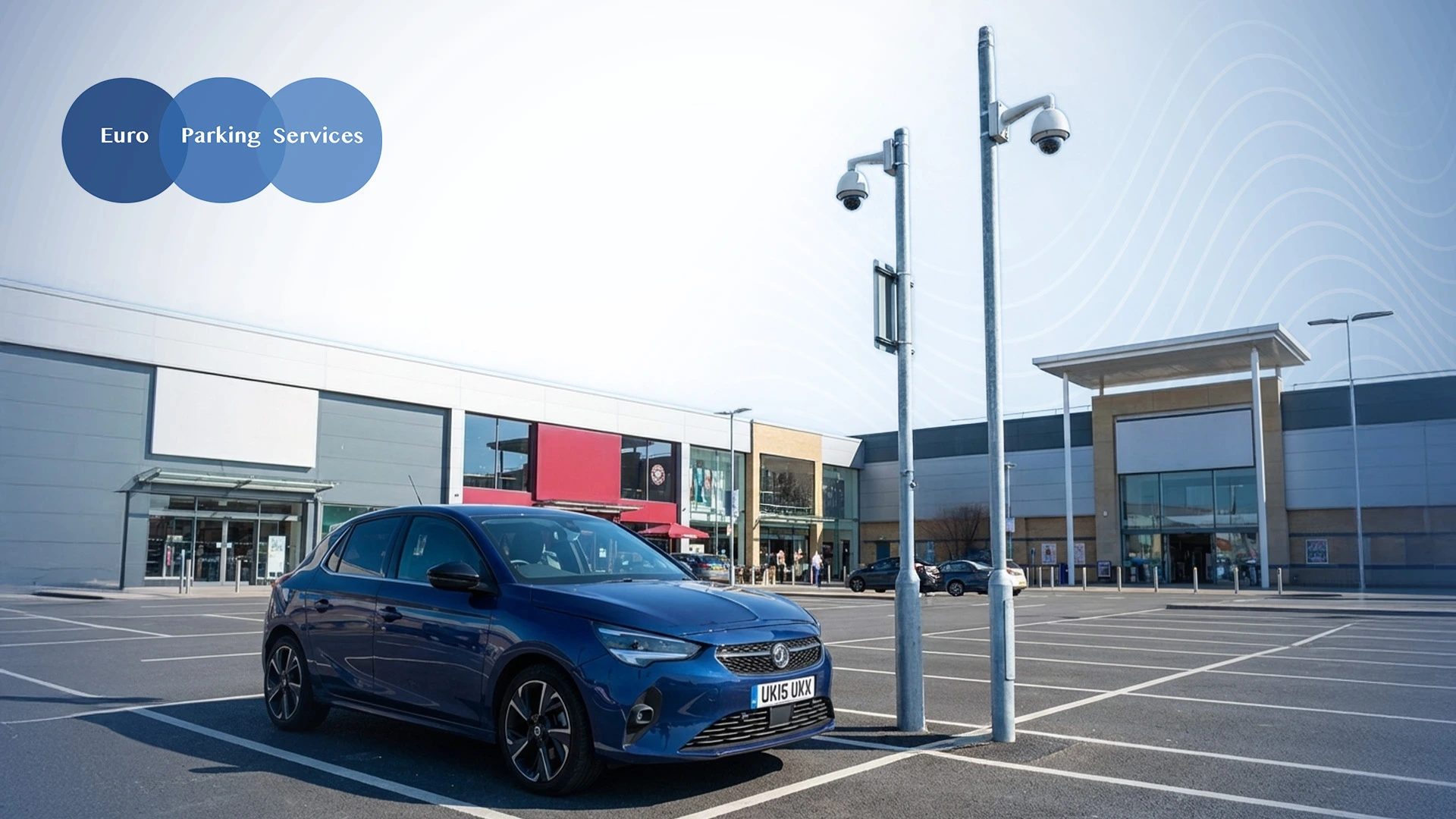 Blue car parked in a shopping center parking space under surveillance cameras.